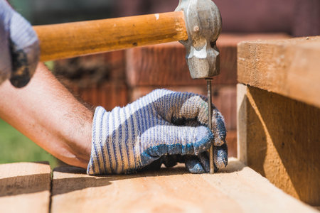 Close-up of a nail being driven into a board with a hammer. A carpenter wearing cloth protective gloves is nailing wooden boards.の写真素材