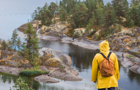 Tourism. Hiking. Ladoga skerries on Lake Ladoga. A tourist man in a yellow raincoat with a backpack on the rocky islands looks into the distance. Republic of Karelia, Russia.の写真素材