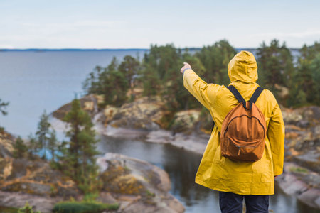 Tourism. Hiking. Ladoga skerries on Lake Ladoga. A tourist man in a yellow raincoat with a backpack on the rocky islands looks into the distance. Republic of Karelia, Russia.の写真素材