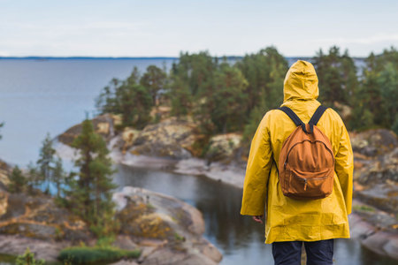 Tourism. Hiking. Ladoga skerries on Lake Ladoga. A tourist man in a yellow raincoat with a backpack on the rocky islands looks into the distance. Republic of Karelia, Russia.の写真素材