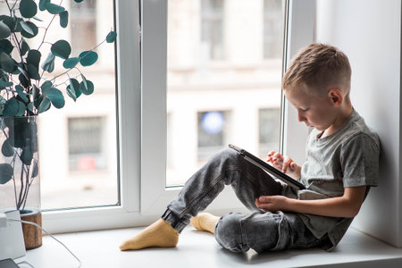 A 6-year-old boy is sitting on a windowsill with a tablet. A child is playing with a phone.の写真素材