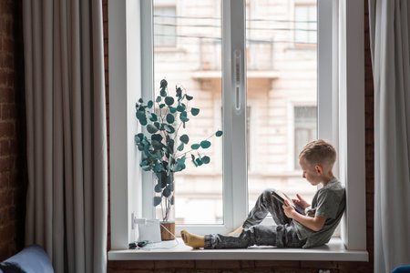A 6-year-old boy is sitting on a windowsill with a tablet. A child is playing with a phone. Children...