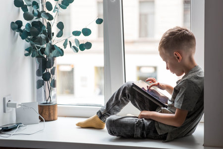 A 6-year-old boy is sitting on a windowsill with a tablet. A child is playing with a phone.の写真素材