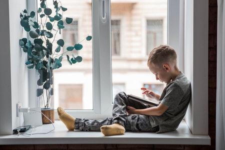 A 6-year-old boy is sitting on a windowsill with a tablet. A child is playing with a phone. Children and gadgets.の写真素材