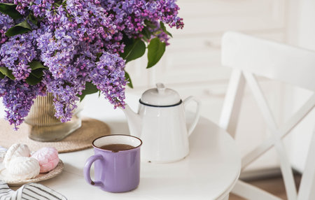 The concept of home comfort. A large purple bouquet of lilacs, a ceramic teapot, a cup and a delicious marshmallow on the table. In the background is the interior of a modern white kitchen.の写真素材