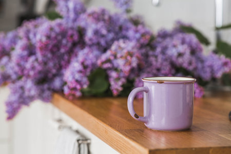 The concept of home comfort and drinks. A lilac cup of tea or coffee on the kitchen countertop. In the background is a large purple bouquet of lilacs.の写真素材