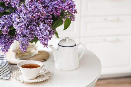 The concept of home comfort. A large purple bouquet of lilacs, a ceramic teapot and a cup on the table. In the background is the interior of a modern white kitchen. The concept of home comfort.の写真素材