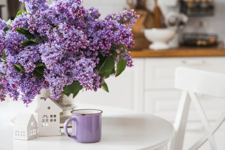 The concept of home comfort. A large purple bouquet of lilacs, white Scandinavian houses and a cup of tea on the table. In the background is the interior of a modern white kitchen.の写真素材