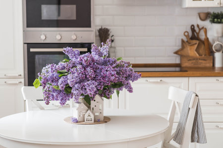 The concept of home comfort. A large purple bouquet of lilacs and white Scandinavian houses on the table. In the background is the interior of a modern white kitchen.の写真素材
