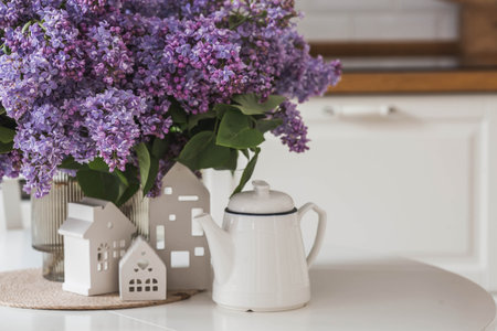The concept of home comfort. A large purple bouquet of lilacs, white Scandinavian houses and a ceramic teapot on the table. In the background is the interior of a modern white kitchen.の写真素材