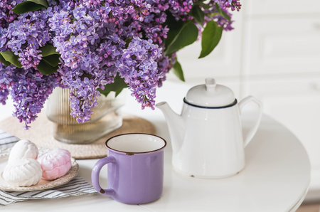 The concept of home comfort. A large purple bouquet of lilacs, a ceramic teapot, a cup and a delicious marshmallow on the table. In the background is the interior of a modern white kitchen.の写真素材