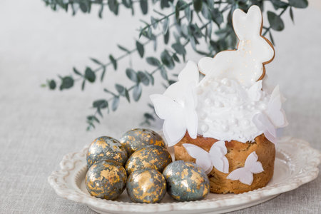 Easter cake with a white rabbit and butterflies, Easter eggs with gold and eucalyptus branches on a white kitchen table. Stylish Easter still life. Happy Easter 2026.の写真素材