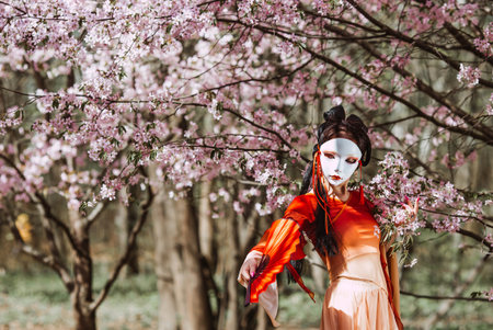 The portrait symbolizes the traditions and culture of the Japanese people. A woman wearing a Japanese mask on her face, traditional clothing, and a fan in her hand, surrounded by cherry.の写真素材