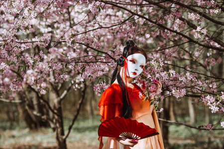 The portrait symbolizes the traditions and culture of the Japanese people. A woman wearing a Japanese mask on her face, traditional clothing, and a fan in her hand, surrounded by cherry.の写真素材