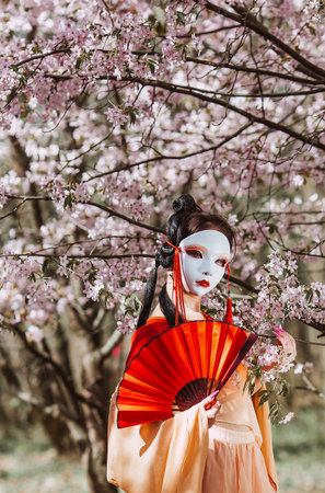 The portrait symbolizes the traditions and culture of the Japanese people. A woman wearing a Japanese mask on her face, traditional clothing, and a fan in her hand, surrounded by cherry.の写真素材