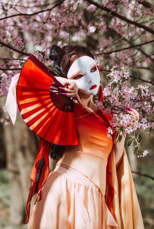 The portrait symbolizes the traditions and culture of the Japanese people. A woman wearing a Japanese mask on her face, traditional clothing, and a fan in her hand, surrounded by cherry.の写真素材