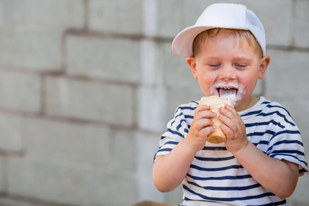 A funny, emotional, happy little boy with a dirty face is eating ice cream on a hot summer day. The child is enjoying a sweet dessert during the summer holidays.の写真素材