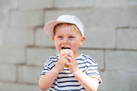 A funny, emotional, happy little boy with a dirty face is eating ice cream on a hot summer day. The child is enjoying a sweet dessert during the summer holidays.の写真素材