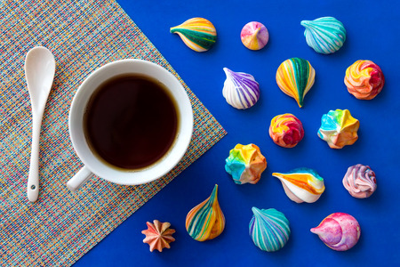 Still life with a cup of tea and a scattering of colorful meringue cakes. Appetizing meringues. Background of a multi-colored mat and bright blue base.の写真素材
