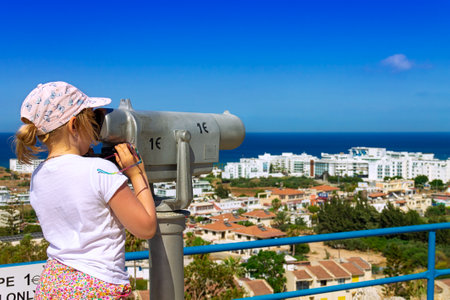 Cyprus, Protaras - 22 June 2021. A girl looks through binoculars at the village and the Mediterranean Sea from the mountain on top of which is the Church of Saint Elias. Bright midday sun.のeditorial素材