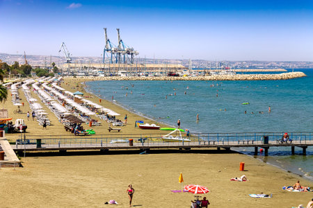 Cyprus, Larnaca - 28 June 2021. Finikoudes beach. The seaport is visible in the distance. People walk along the pier, sunbathe and swim in the sea.のeditorial素材