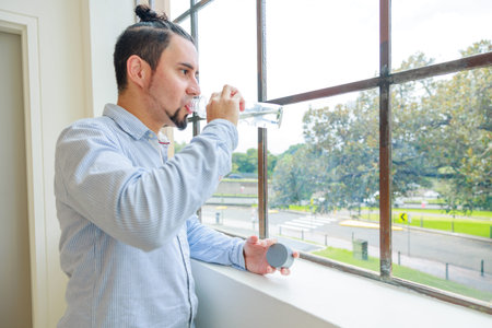 Man drinking water from a bottle while standing near the window at home