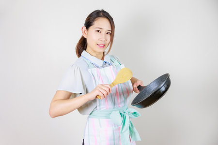 Portrait of Asian housewife in apron holding frying pan and spatulaの写真素材