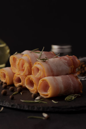 Rolls of bacon with rosemary and peppercorns on black backgroundの写真素材