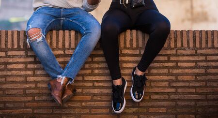 Couple sitting on brick roman wall undefined. Men in blue jeans and brown boots, woman in black leggings and sneakers, orange wall as a benchの写真素材