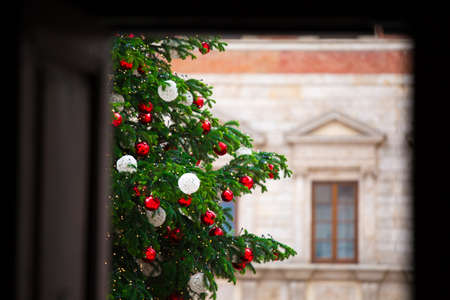 Window view on emerald green Xmas tree with light garlands, white and red balls. Medieval house with in background out of focus. New Year. Isolation at home. Quarantine. Live spruce.の写真素材