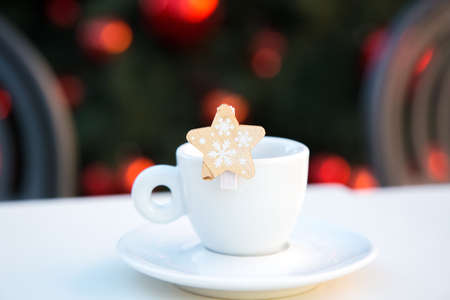 White porcelain coffee cup with saucer and wooden decorative star with snowflakes. Blurred background with chairs and Christmas tree with red balls. New year bar scene. Congratulations. Celebrating alone. High quality photoの写真素材