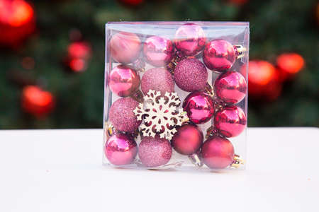 Plastic transparent square box with rose and purple small Christmas balls and wooden snowflake inside. White table. New year tree with red decorations in background. Xmas toys. Preparation for holidays. High quality photoの写真素材