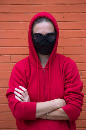 Woman looks straight in camera. Red sport hoodie with hood put on head in black sunglasses and black protective medical mask. Underground style in covid world. Orange brick wall behind. Caucasian girlの写真素材