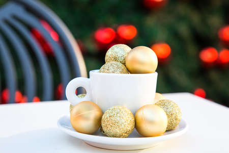 Coffee cup with golden Christmas balls inside. Coffee pair on white table, grey chair and new year tree with red toys in background out of focus. Winter cafe menu. Post card picture. Party table setting. High quality photoの写真素材