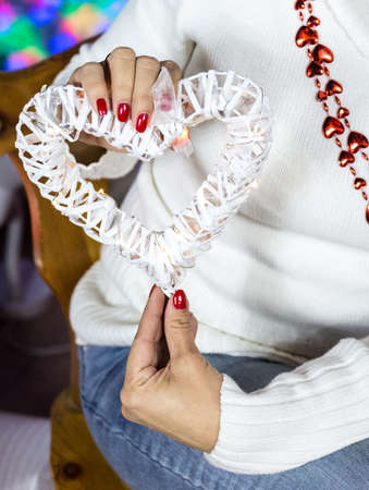 Vertical picture of woman in white sweater, holds in hands white wooden heart with electric garland. New year decoration. Fingers with red nails. Tender and love concept. Saint Valentains dayの写真素材