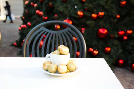 Coffee cup with golden Christmas balls inside. Coffee pair on white table, grey chair and new year tree with red toys in background out of focus. Winter cafe menu. Post card picture. Party table setting. High quality photoの写真素材