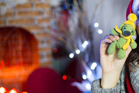 Female hand holds small green teddy bear in yellow hat and scarf. Blurred fireplace with new year lights and decorations. Hand made knitted toy as Christmas present. Preparing gifts. postcard pictureの写真素材