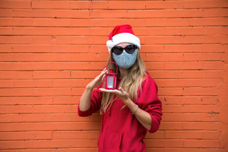 Blond girl in silver mask, red hat and black sun glasses holds in hands Christmas light. Caucasian woman in Christmas costume and covid protection. Orange brick wall behind, copy space. Reusable maskの写真素材