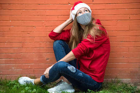 Caucasian blond girl in red hat, coat, jeans and silver protective mask sits on green grass with crossed legs and looks up. Preparation for Christmas 2020. Celebrating pandemic new 2021 year.の写真素材