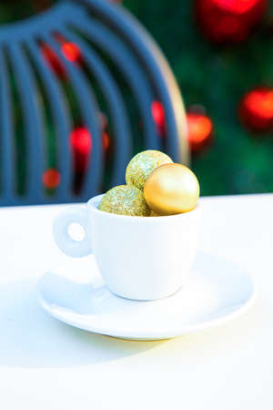 Coffee cup with golden Christmas balls inside. Coffee pair on white table, grey chair and new year tree with red toys in background out of focus. Winter cafe menu.Copy space. Post card picture. Party table setting. High quality photoの写真素材