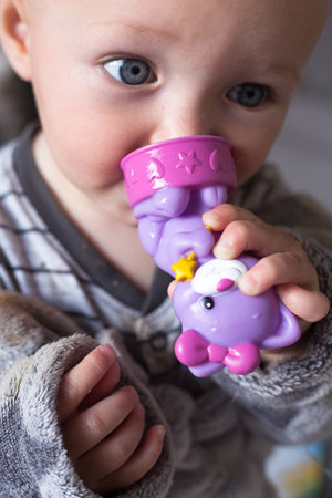 Blue eye baby girl in grey clothes and bathrobe holds in hands and nibble a violet toy cat. First tooth. Closeup portrait of caucasian child. Teething problem. After bath. Tender and emotional conceptの写真素材
