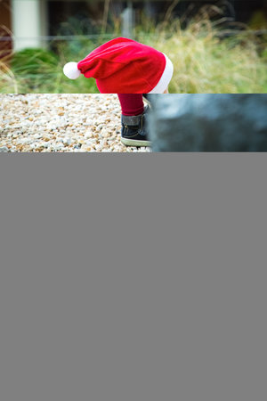 Baby girl dressed for Christmas walks in a park with new year bag. Red hat. dress and tights, blue coat, felt bag in form of winter house. No face. Holiday concept. High quality photoの写真素材