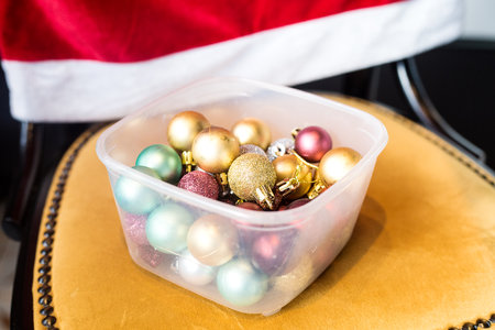 Golden, blue and burgundy Christmas balls in a transparent plastic food container on vintage orange chair with red cover. Storage of new year decoration. Space managementの写真素材