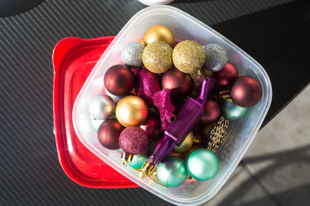 Golden, blue and burgundy Christmas balls packed for keeping in a transparent plastic food container on black table. Red cover open. Top view. Storage of new year decoration. Space managementの写真素材