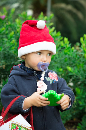 Baby girl dressed for Christmas walks outdoors with green trees behind. Red hat, blue coat, felt bag, green toy in hands. New year tree. Holiday concept. Day light portrait. Caucasian blue eye babyの写真素材