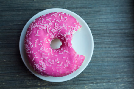 Top view to bright pink donut on white round plate bitten off once. Aged wooden background. Isolated object. Traditional American sweets for breakfast. Copy space. White sugar decoration on top.の写真素材