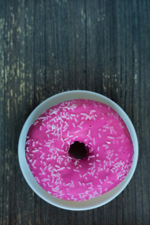 Vertical picture of bright pink donut on white round plate. Aged wooden background. Isolated object close up. Traditional American sweets for breakfast. Copy space. White sugar decoration on top.の写真素材