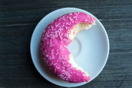 Top view to half eaten bright pink donut on white round plate. Aged wooden background. Isolated object. Traditional American sweets for breakfast. Copy space. White sugar decoration on top.の写真素材