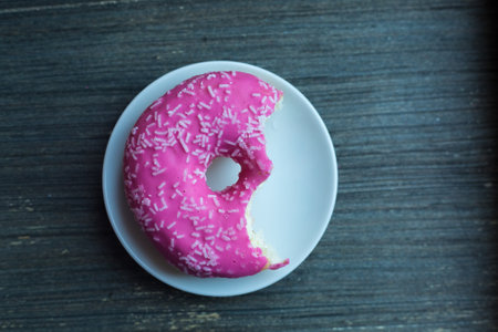 Top view to bright pink donut on white round plate bitten off twice. Aged wooden background. Isolated object. Traditional American sweets for breakfast. Copy space. White sugar decoration on top.の写真素材