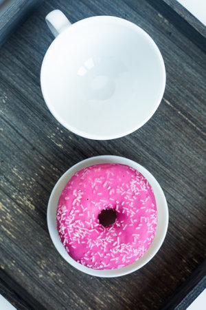 Vertical picture of bright pink donut on white round plate and tea cup above. Aged wooden background. Isolated objects. Traditional American breakfast. Copy space. Geometrical concept. Eating at homeの写真素材